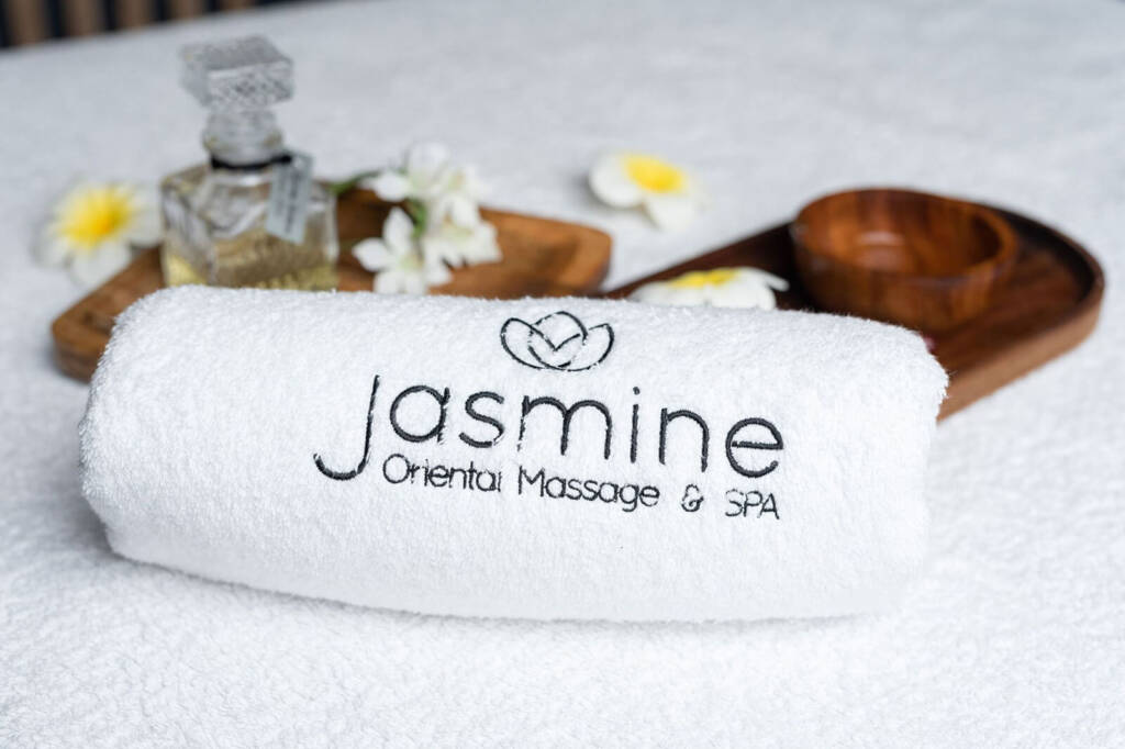Zrolowany biały ręcznik z wyhaftowanym napisem "Jasmine Oriental Massage & SPA", umieszczony na białej powierzchni z drewnianą tacą, szklaną butelką i kwiatami w tle.